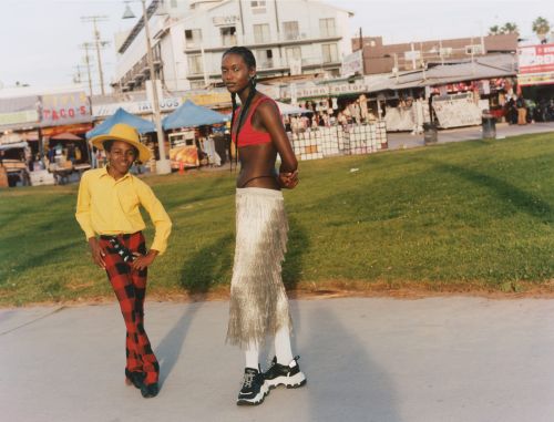 Two fashionably dressed people posing outdoors on a sunny day. One is wearing a yellow shirt and red checkered pants, the other a red top and white skirt.