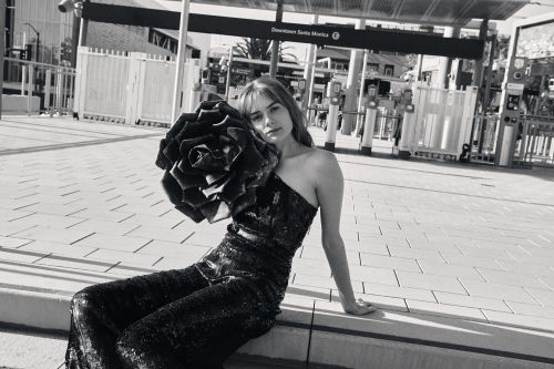 Black and white fashion portrait of a woman in a black one-shoulder dress with a large rose detail, sitting in front of a transit station.