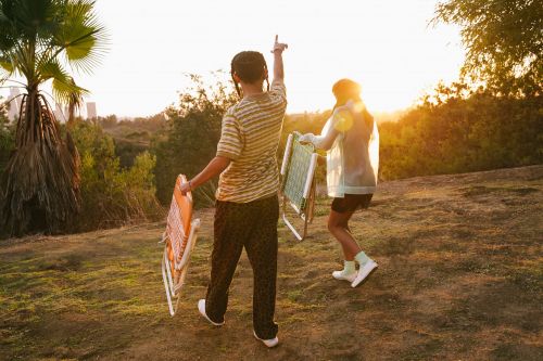 Two people walk across a grassy field carrying folding chairs at golden hour, silhouetted against a treeline and distant buildings.