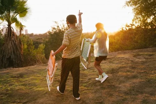Two people walk across a grassy field carrying folding chairs at golden hour, silhouetted against a treeline and distant buildings.