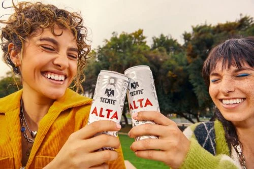 Two smiling women outdoors toasting cans of Tecate Alta beer in a medium shot, against a backdrop of trees and overcast sky.