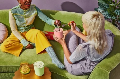 Two smiling women sit on a green couch, sharing ice cream from a pink container. There are beverages and a plant nearby indoors.