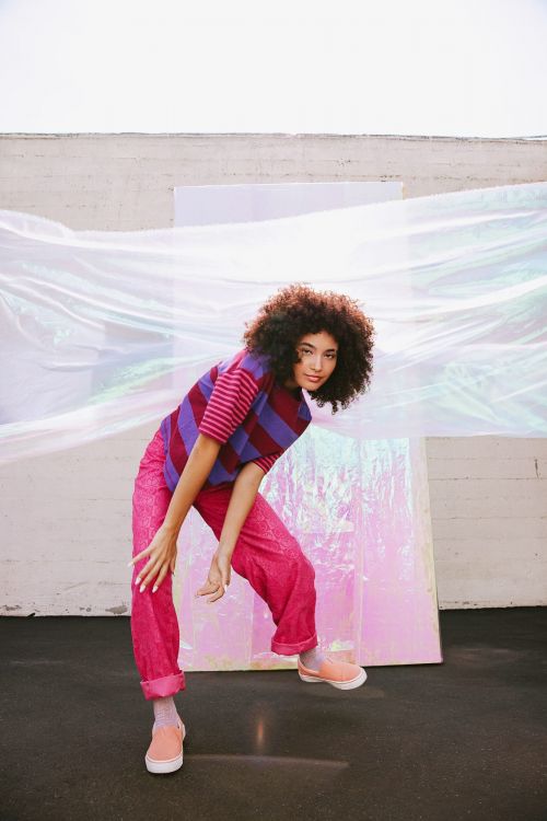 A woman with curly hair poses in a striped shirt and pink pants against a white wall and iridescent sheet, with a playful expression.