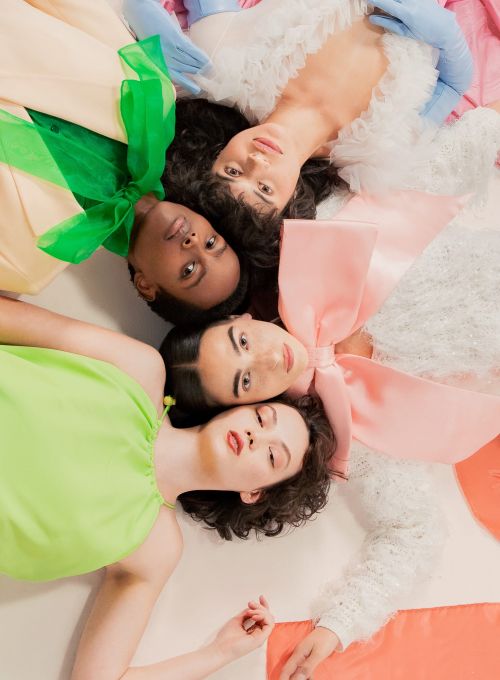 Overhead shot of four diverse women lying in a circle wearing pastel-colored dresses, featuring decorative bows, against a white background.