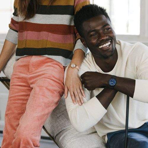 Couple in relaxed indoor setting showcasing wristwatches; man smiles, woman in striped sweater and corduroy pants stands beside him.