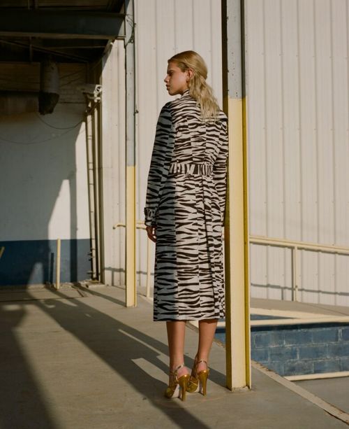 A blonde woman poses on a walkway wearing a tiger-striped trench coat, gold heels, and a half-up hairstyle near a white building.