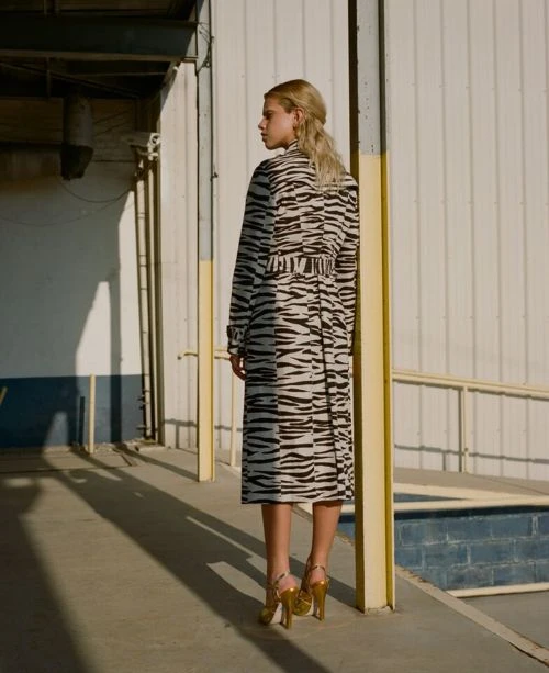 A blonde woman poses on a walkway wearing a tiger-striped trench coat, gold heels, and a half-up hairstyle near a white building.