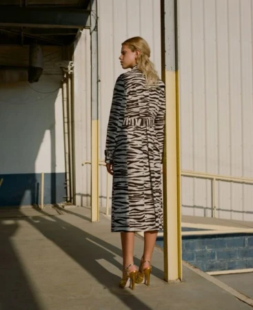 A blonde woman poses on a walkway wearing a tiger-striped trench coat, gold heels, and a half-up hairstyle near a white building.