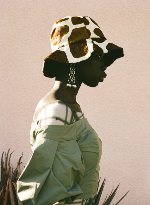 Profile shot of an adult woman with dark skin in an off-shoulder top and spotted bucket hat, standing against a pale pink wall.
