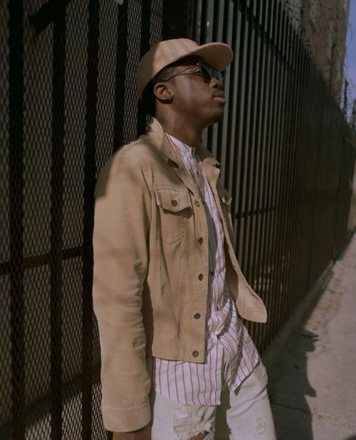 Man in tan jacket, baseball cap, and sunglasses leaning against black metal fence under bright sun, in an urban outdoor environment.