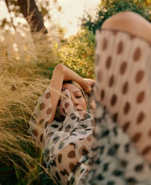 Woman in sheer polka dot top and light hair rests in tall golden grass in natural light, outdoor portrait.