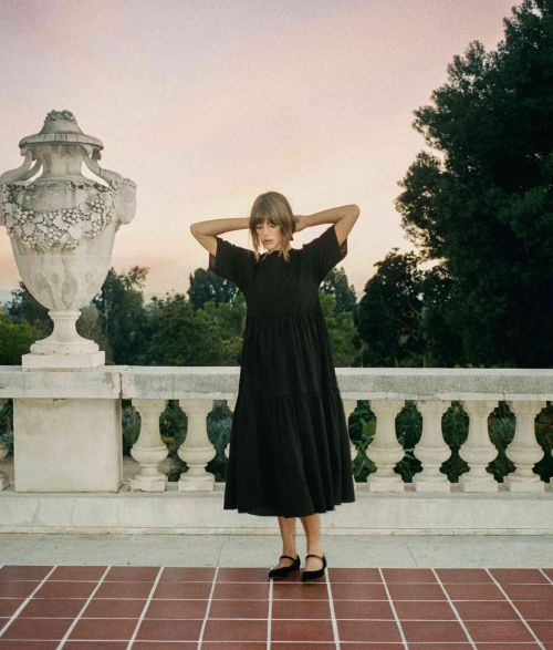 Adult female in a black dress standing on a tiled patio in front of a white stone railing and foliage with a golden sky at sunset.
