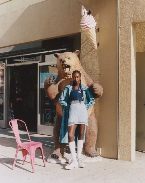 An adult woman poses in front of a bear statue and an ice cream cone sign. The outdoor scene features an urban setting with natural light.