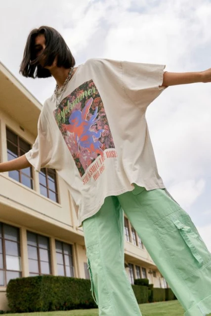 Woman wearing a white t-shirt with graphic and mint-green cargo pants stands outside a light-beige building on a sunny day.