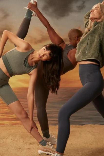 Four diverse women in athletic wear stretch on a sandy beach with a soft, blurred ocean and sky background during golden hour.