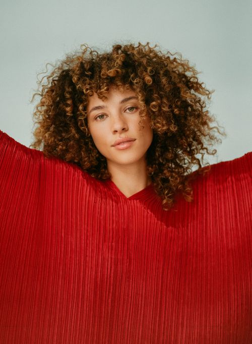 Portrait of a woman with curly brown hair wearing a pleated red top against a light grey background