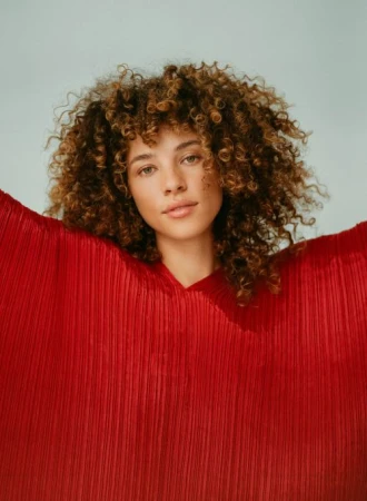 Portrait of a woman with curly brown hair wearing a pleated red top against a light grey background