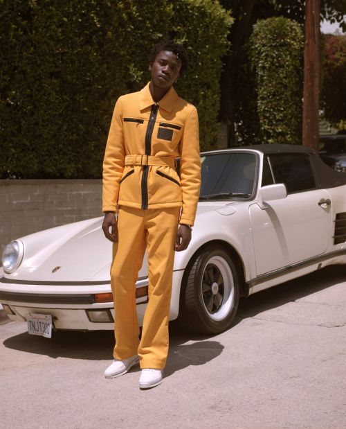 An adult Black male model poses next to a white convertible sports car outside wearing a yellow-and-black matching suit.