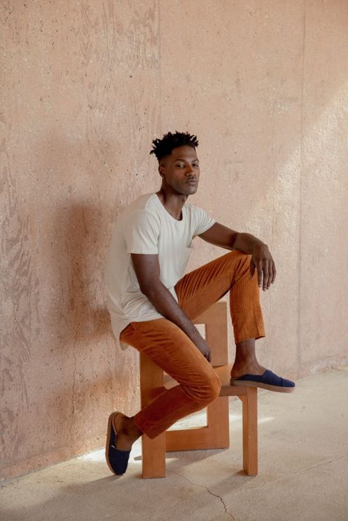 An adult African-American man wearing a tee shirt and slacks poses indoors, sitting on a stool, in a lifestyle fashion shot against a textured wall.