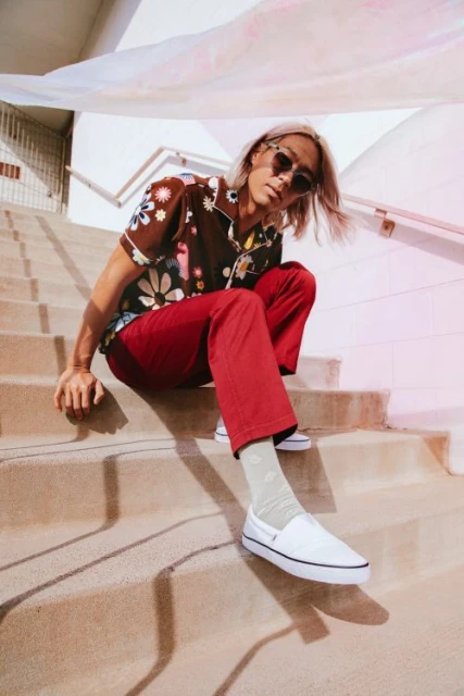 Man sits on outdoor stairs wearing sunglasses, floral shirt, red pants, white slip-on shoes, light socks, under bright sunlight.
