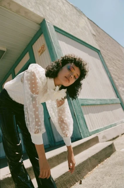 A curly-haired woman in a sheer polka-dot blouse and shiny black pants strikes a pose against a light blue-trimmed building exterior.