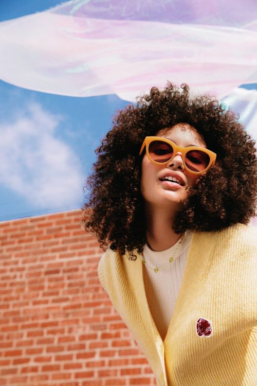 Stylish woman with curly hair poses in a yellow cardigan and sunglasses in front of a brick wall and blue sky.