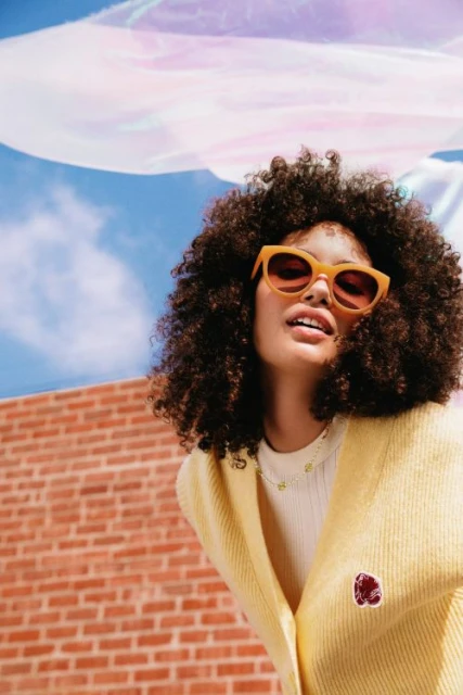 Stylish woman with curly hair poses in a yellow cardigan and sunglasses in front of a brick wall and blue sky.