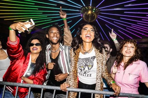Group of stylish young adults at a nightclub with a disco ball, rainbow neon lights, and metal crowd barrier enjoying nightlife.