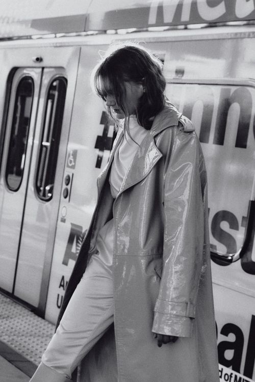 Woman in a vinyl trench coat stands on a train platform in black and white. The train is visible in the background.