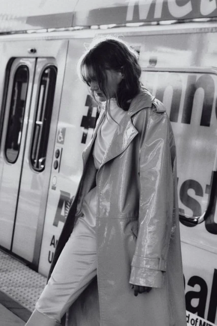 Woman in a vinyl trench coat stands on a train platform in black and white. The train is visible in the background.