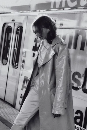 Woman in a vinyl trench coat stands on a train platform in black and white. The train is visible in the background.