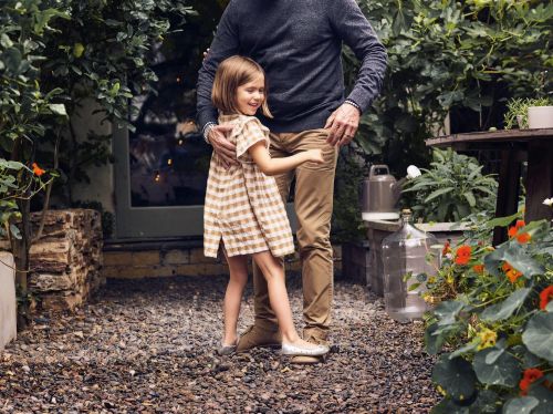 A young girl in a checkered dress stands outside with an adult, surrounded by plants and flowers in a garden setting with natural light.
