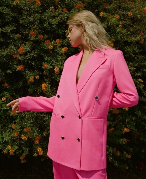 A young adult woman models a bright pink, double-breasted blazer and matching pants outdoors with a flowering bush backdrop.