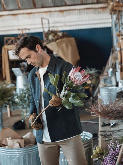 Adult male florist trimming a pink protea flower in a workshop, wearing work gloves, with visible floral and craft supplies.