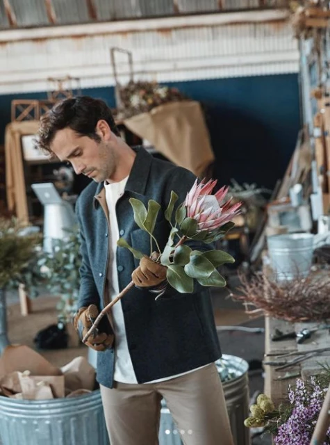 Adult male florist trimming a pink protea flower in a workshop, wearing work gloves, with visible floral and craft supplies.