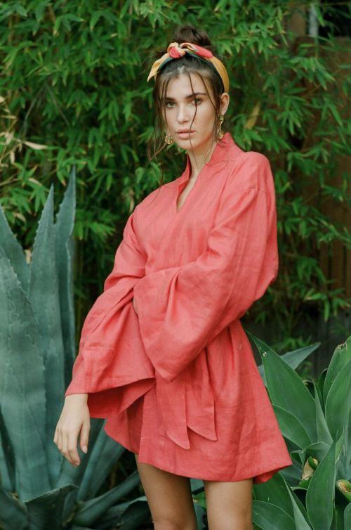 A woman with a hair scarf stands outside wearing a pink kimono-style dress with wide sleeves against a leafy green background.