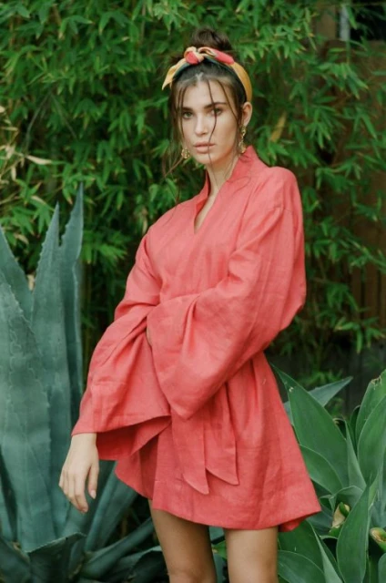 A woman with a hair scarf stands outside wearing a pink kimono-style dress with wide sleeves against a leafy green background.