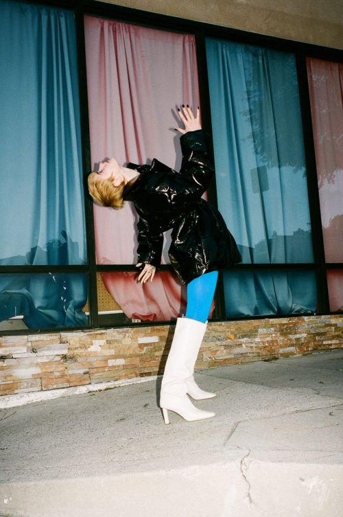 A model poses outdoors wearing a shiny black coat, turquoise leggings, and white heeled boots against a windowed building facade.