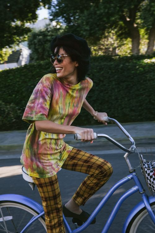 Adult female smiling and riding a blue bicycle outside. She wears a tie-dye shirt, plaid pants, black shoes, a hat, and sunglasses.