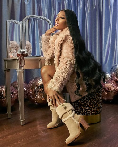 Adult female in a fur coat and boots sits at a vanity with makeup and mirror in a studio with disco balls and blue curtains.
