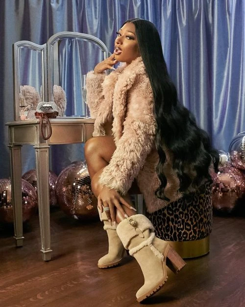 Adult female in a fur coat and boots sits at a vanity with makeup and mirror in a studio with disco balls and blue curtains.