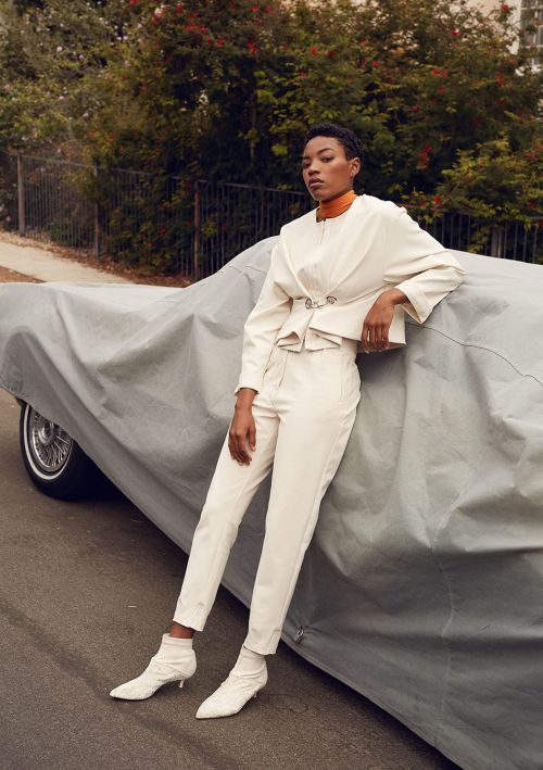 A stylish young adult woman in a white pantsuit poses against a car covered with a gray car cover on a tree-lined street.