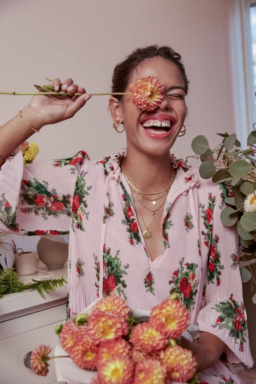 Cheerful woman in a floral dress laughs with a dahlia flower covering one eye in an indoor setting with a bright window in the background.