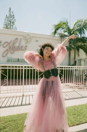 Woman poses outside in a pink tulle dress and black corset with palm trees and a restaurant in the background on a sunny day.