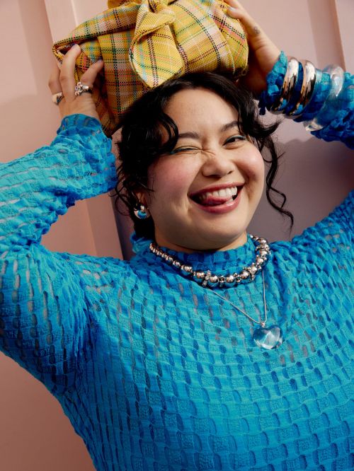 A woman poses indoors, wearing a blue textured top and jewelry, with a yellow plaid-wrapped object on her head, winking at the camera.