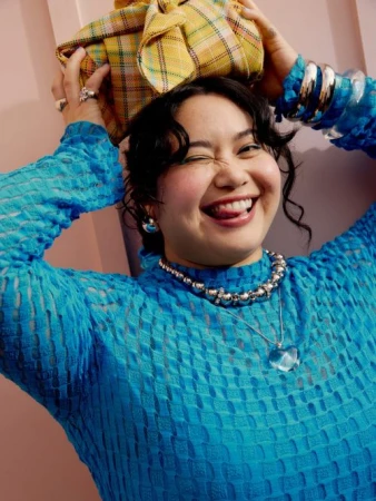 A woman poses indoors, wearing a blue textured top and jewelry, with a yellow plaid-wrapped object on her head, winking at the camera.