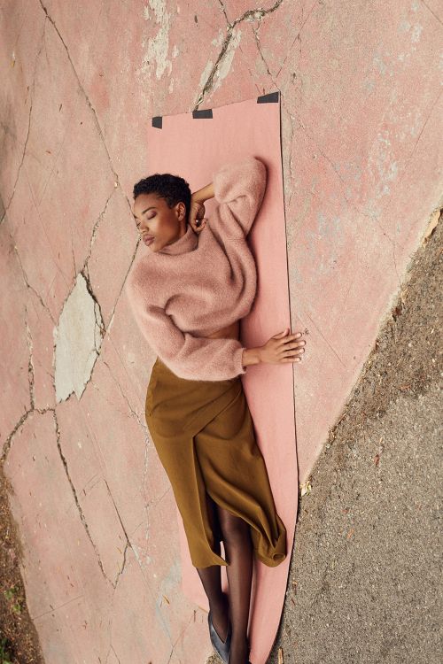 Overhead shot of a woman in a pink sweater and brown skirt laying on a pink mat on a cracked pink and grey concrete surface.