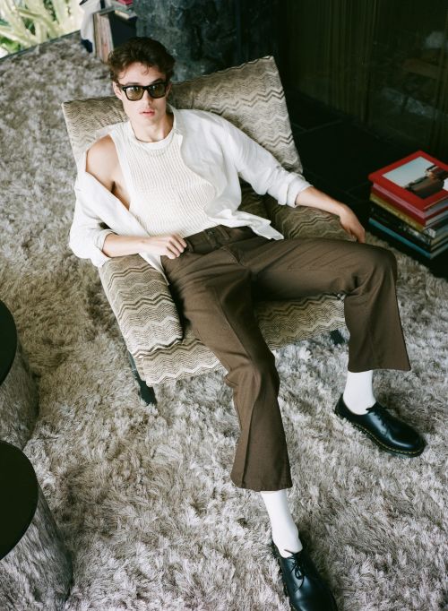 Male fashion model poses in a patterned armchair in a sunlit room with stacks of books, wearing a white top, brown trousers, and black shoes.