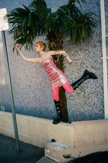 A woman in a pink dress and boots leaps from a low wall near a palm tree, illuminated by daylight, capturing a fashion-forward moment.
