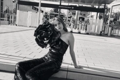 Woman in sequined one-shoulder jumpsuit holding a large decorative rose sculpture in Los Angeles.